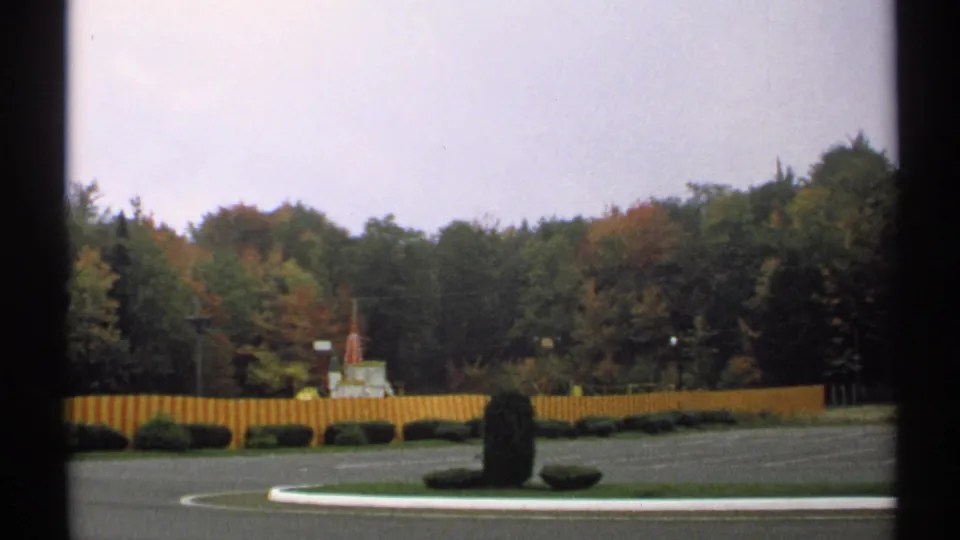 1971 New York home-movie frame of a quiet roadside or park approach with a fence line, shrubs, and autumn-toned trees.