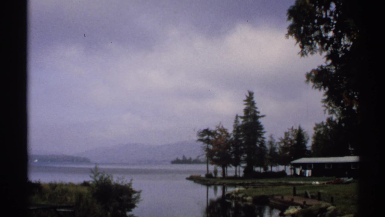 1971 New York 8mm view across a misty lake toward trees, low hills, a small shoreline cabin, and a dock under heavy clouds.