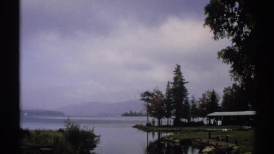 1971 New York 8mm view across a misty lake toward trees, low hills, a small shoreline cabin, and a dock under heavy clouds.