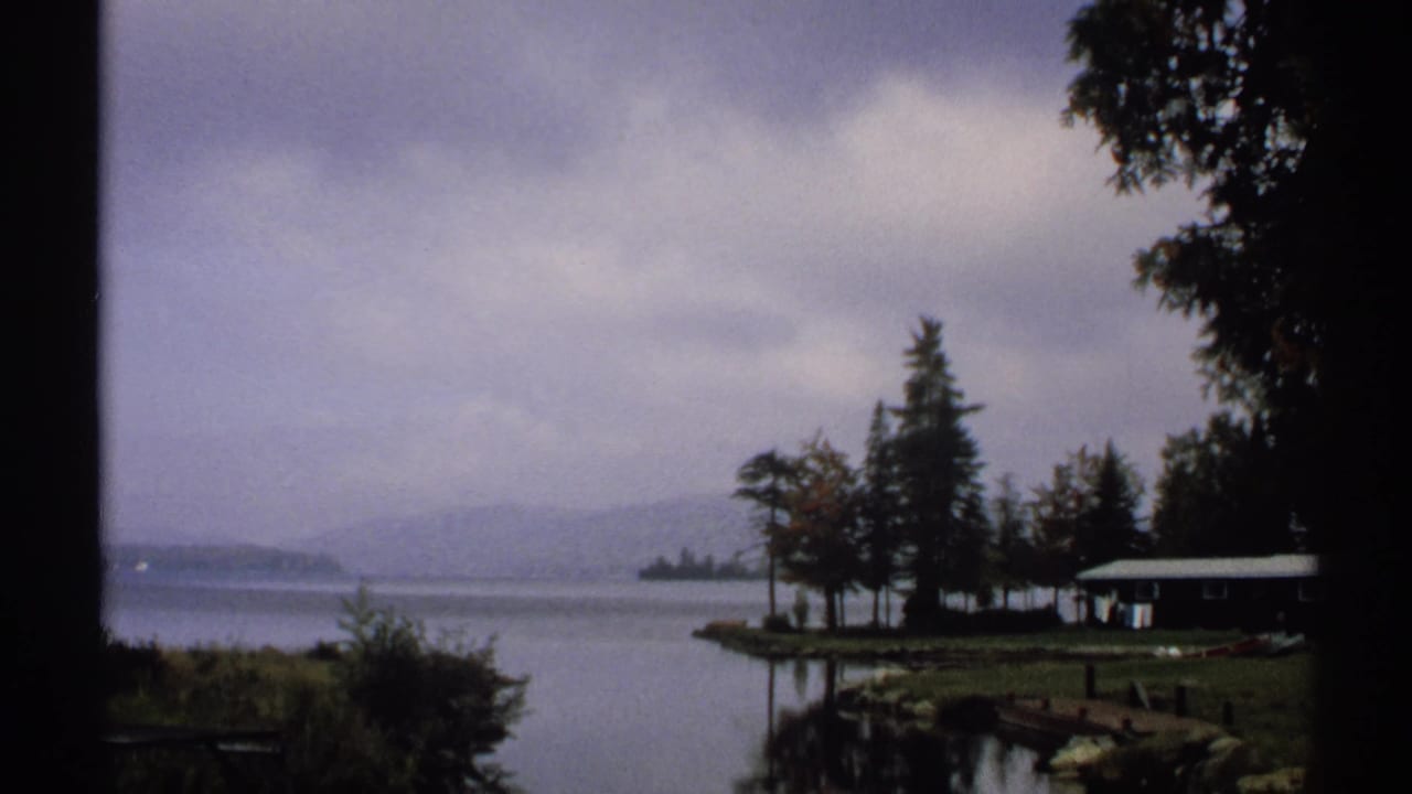 1971 New York home-movie frame of a quiet lakeshore with a small cabin, dock, trees, misty water, and distant hills.