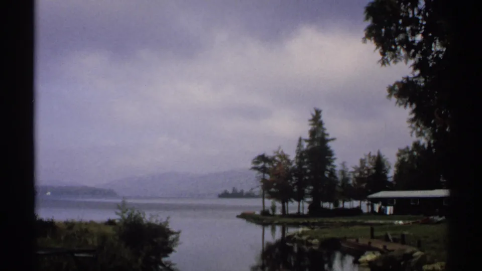 1971 New York home-movie frame of a quiet lakeshore with a small cabin, dock, trees, misty water, and distant hills.