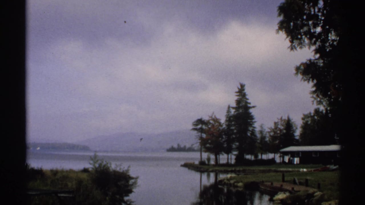 Calm lake with trees, a small cabin, distant hills, and overcast sky.