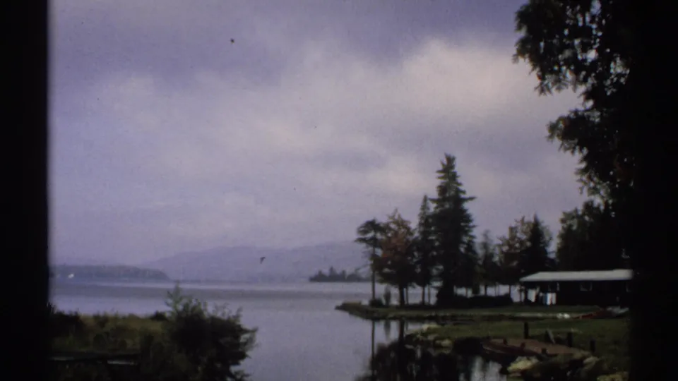 1971 New York 8mm view of a quiet lakeside cabin, tree-lined shore, still water, and hazy hills under cloudy sky.