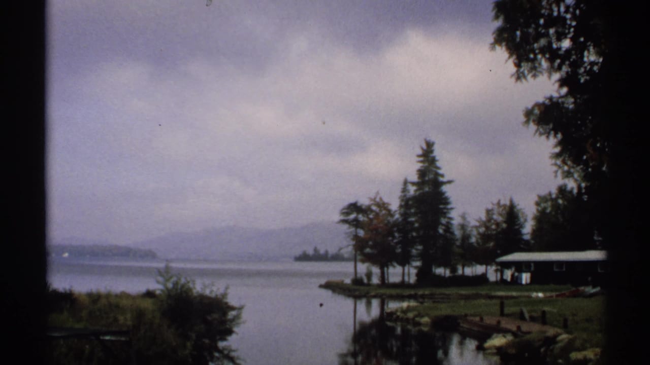 1971 New York home-movie frame showing still lake water, a wooded point, a small cabin, and distant hills in soft overcast light.