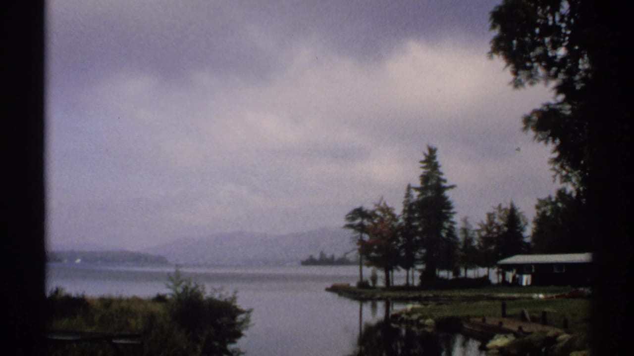 Wide 1971 New York 8mm lakeside view with cloudy sky, calm water, shoreline trees, a small cabin, and softened hills beyond.