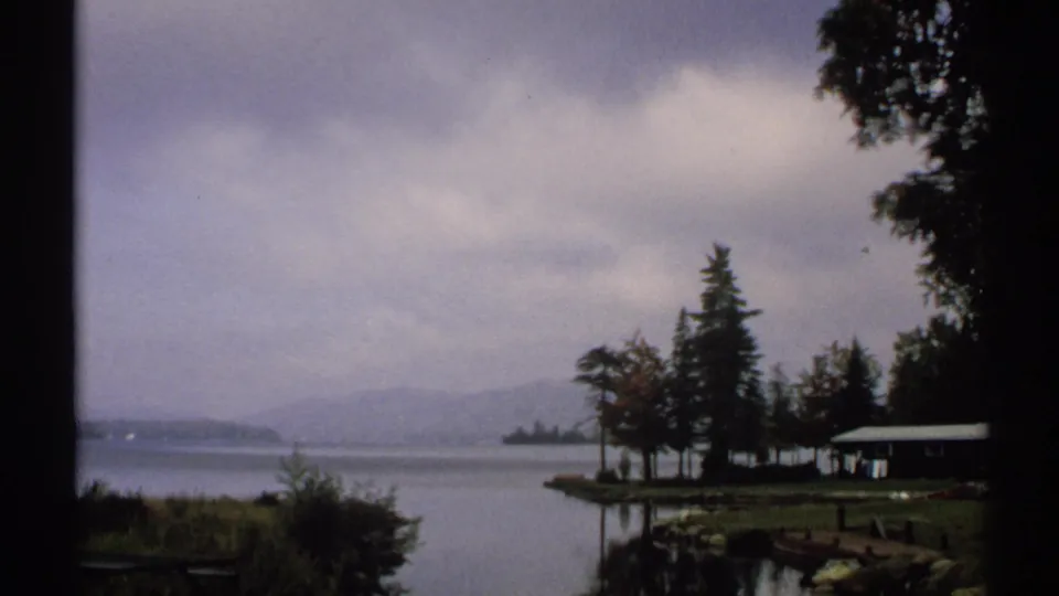 Wide 1971 New York 8mm lakeside view with cloudy sky, calm water, shoreline trees, a small cabin, and softened hills beyond.