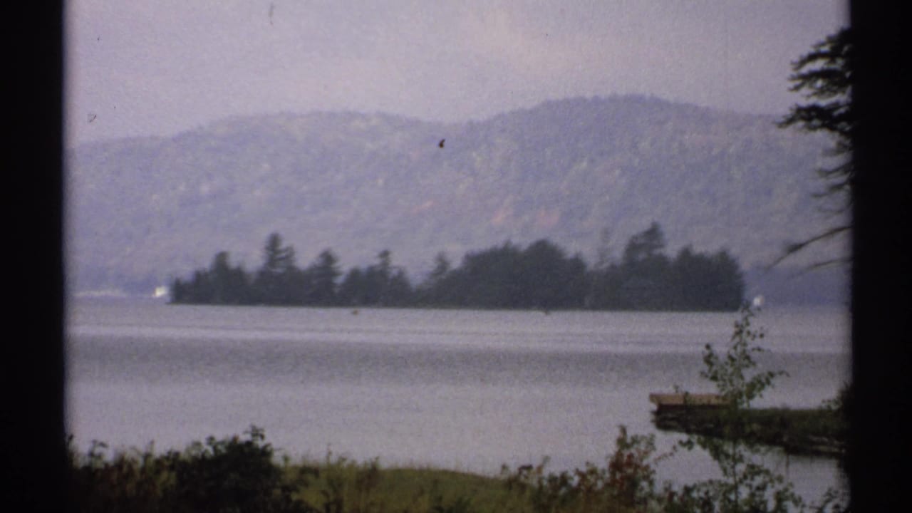 1971 New York 8mm frame of a small island across calm lake water, with hazy hills behind and a dock or shoreline edge at right.