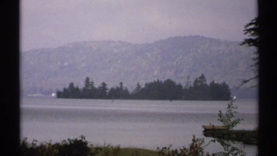 1971 New York 8mm lake view with a wooded island, hazy hills, shoreline plants, and a small dock or platform at right.