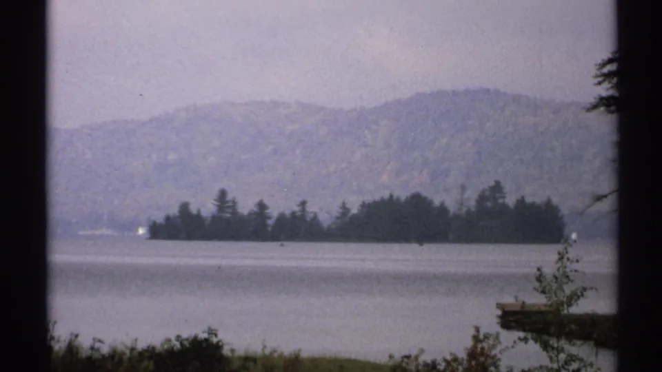 1971 New York home-movie frame of a calm lake, wooded island, hazy hills, and shoreline plants seen through dark frame edges.