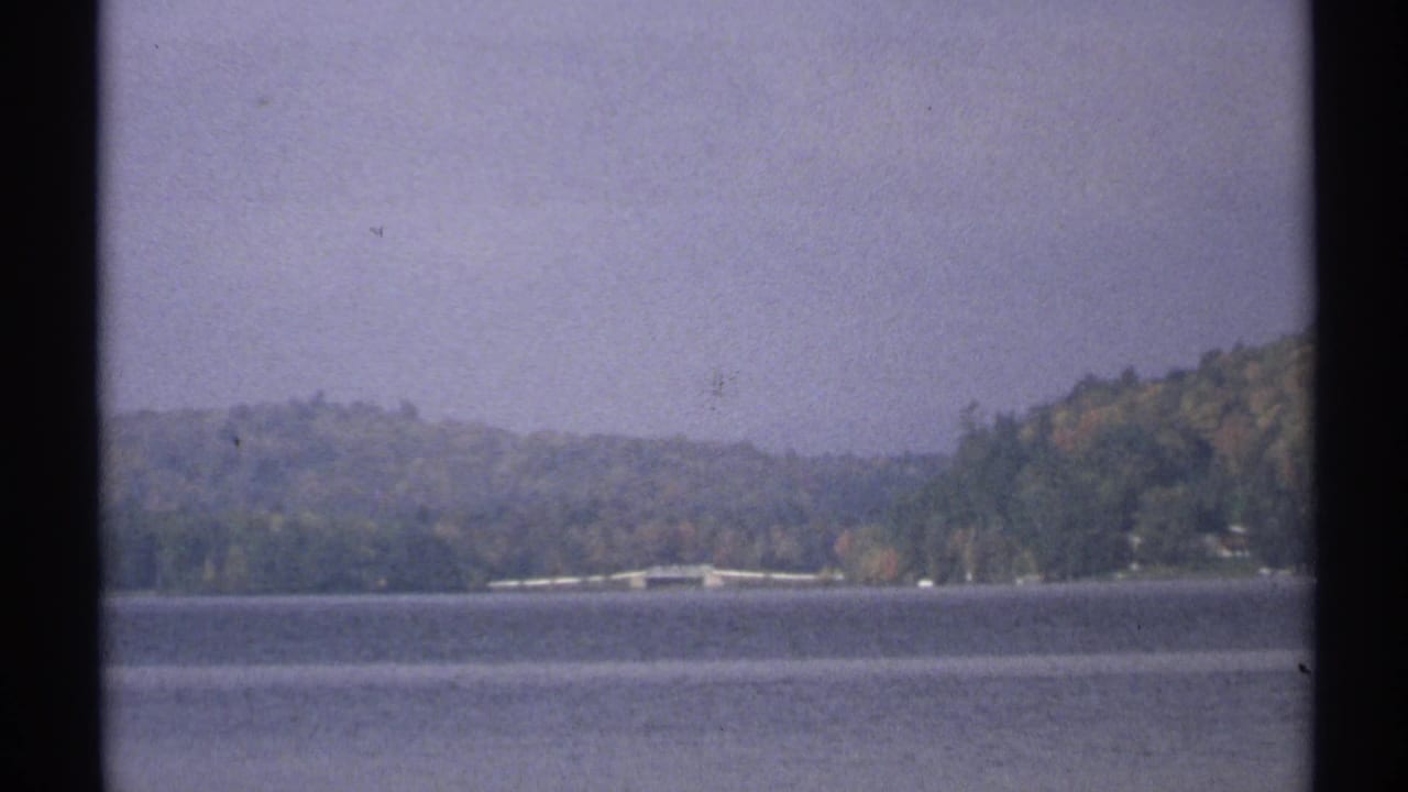 1971 New York 8mm frame showing a broad lake, distant wooded hills, shoreline structures, and an overcast sky.