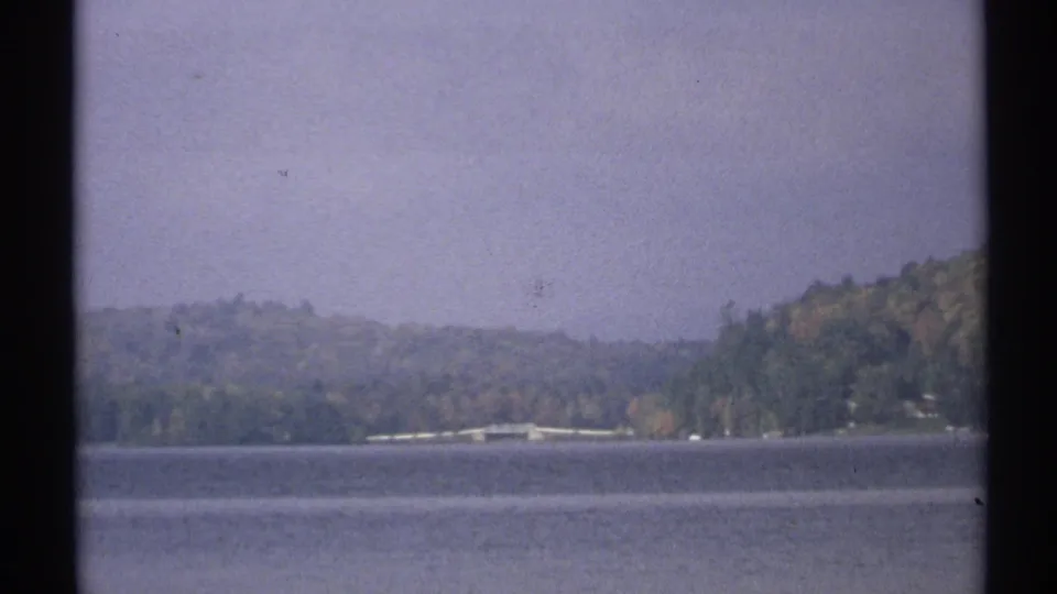 1971 New York 8mm frame showing a broad lake, distant wooded hills, shoreline structures, and an overcast sky.
