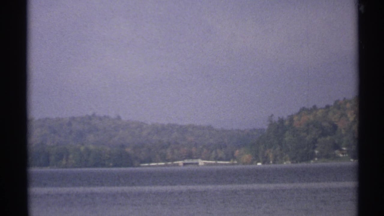 1971 New York home-movie frame of a lake bordered by wooded hills, with a faint shoreline and cloudy sky.