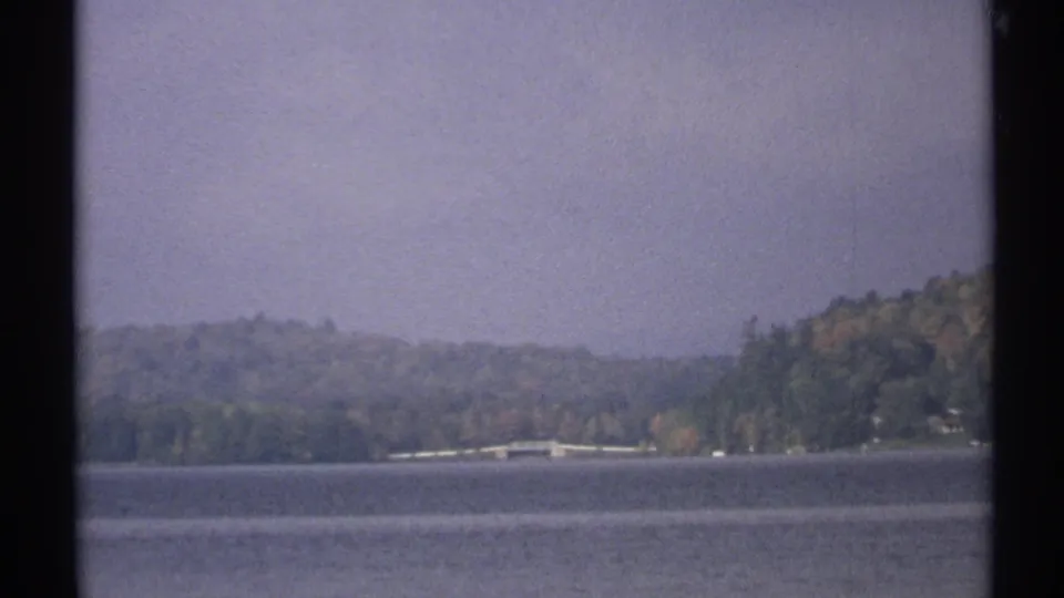 1971 New York home-movie frame of a lake bordered by wooded hills, with a faint shoreline and cloudy sky.
