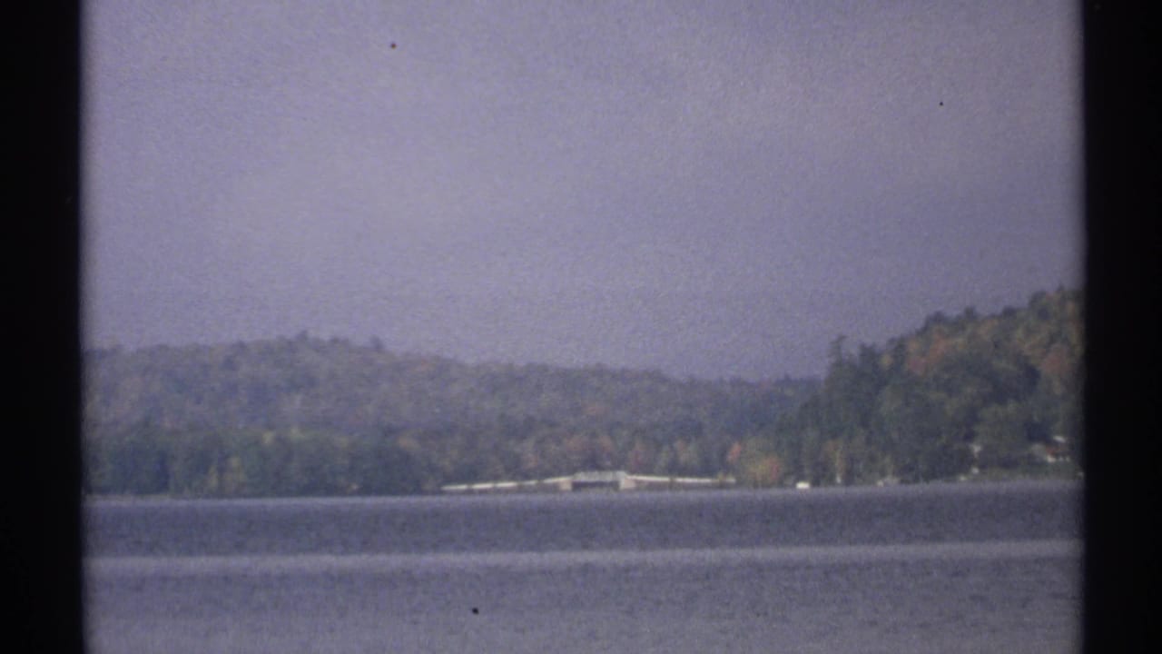 Calm lake with wooded hills and a distant shoreline structure under a hazy sky.