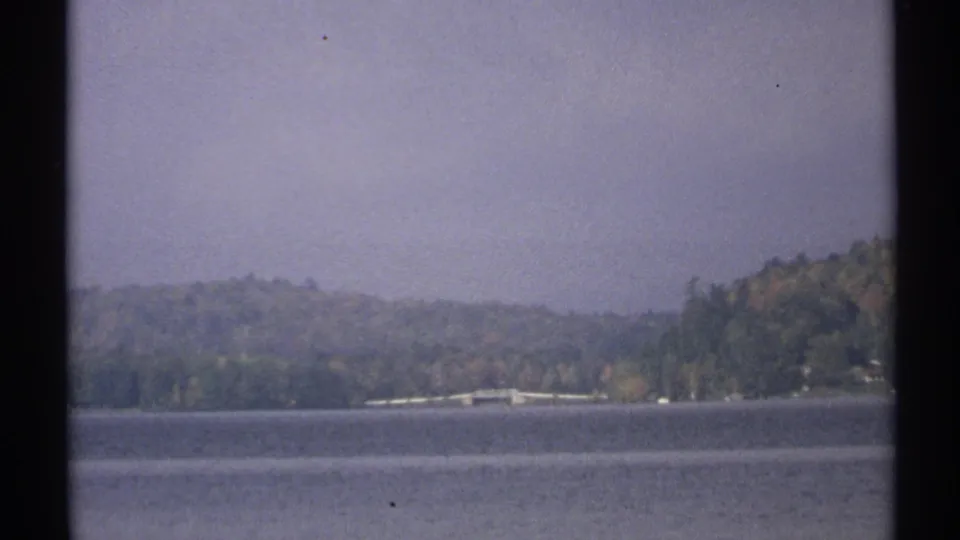 1971 New York 8mm frame showing a calm lake, distant bridge or shoreline structure, and tree-covered hills under haze.