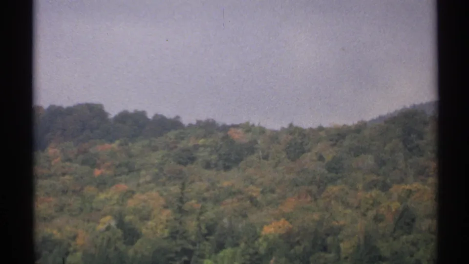 1971 New York 8mm frame showing a distant wooded hillside with green, yellow, and brown fall foliage under a pale sky.