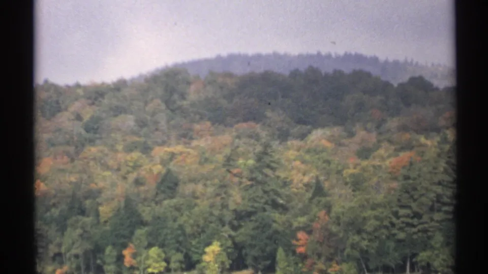 1971 New York 8mm frame showing a wooded hillside with early autumn color beyond still lake water.