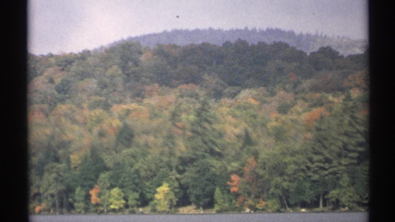1971 New York home-movie view of a forested hill and shoreline reflected near a quiet lake edge.
