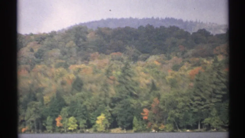 1971 New York home-movie view of a forested hill and shoreline reflected near a quiet lake edge.