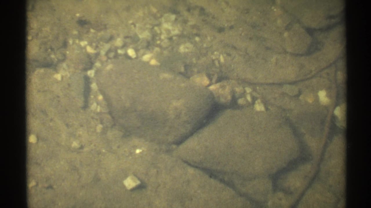 1971 New York 8mm frame looking into clear shallow lake water over smooth stones and pale pebbles.