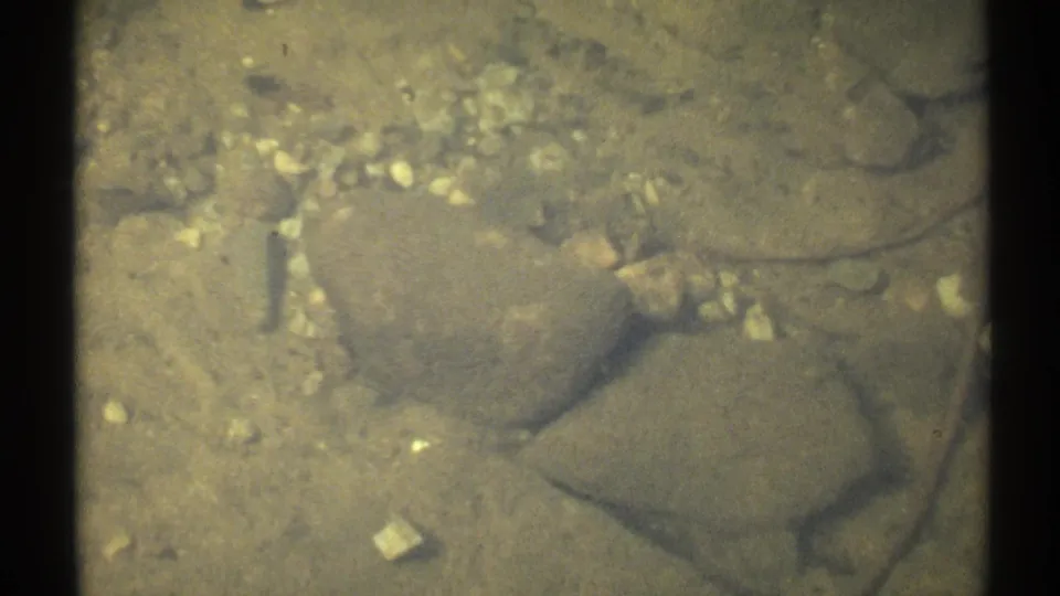 1971 New York 8mm frame showing stones and pebbles beneath shallow water, with soft focus and dark frame edges.