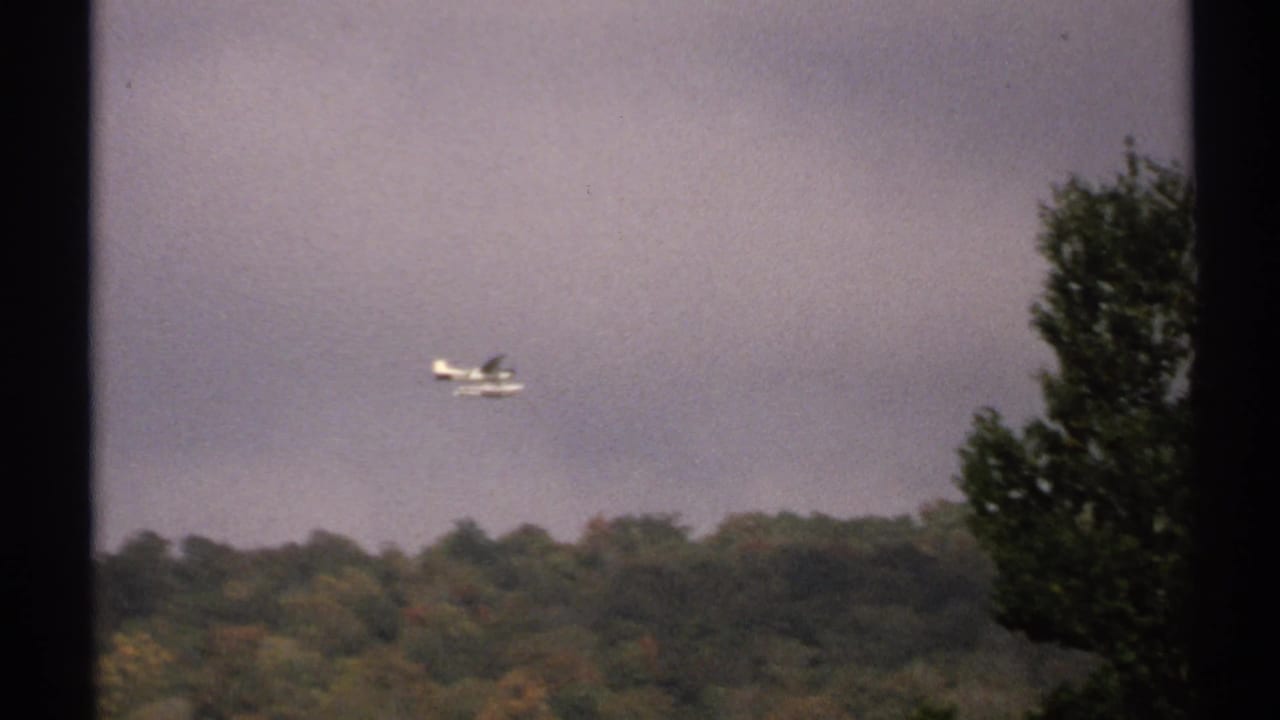 1971 New York 8mm frame showing a distant small seaplane above trees beneath a muted overcast sky.
