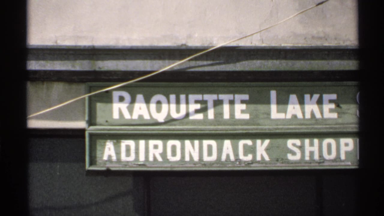 1971 8mm frame of the Raquette Lake Adirondack Shop sign, with white block lettering on a green facade.