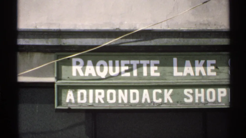 1971 8mm frame of the Raquette Lake Adirondack Shop sign, with white block lettering on a green facade.