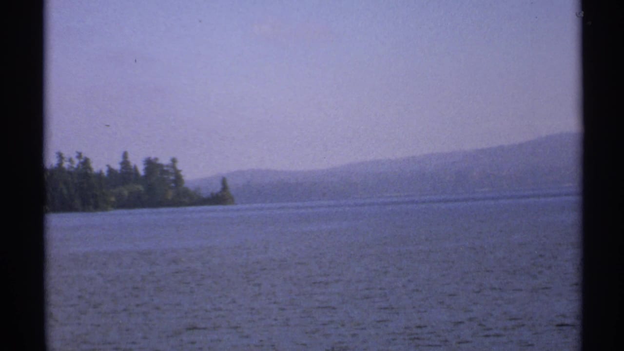 Calm lake water with a wooded shore and hazy hills under a pale sky.