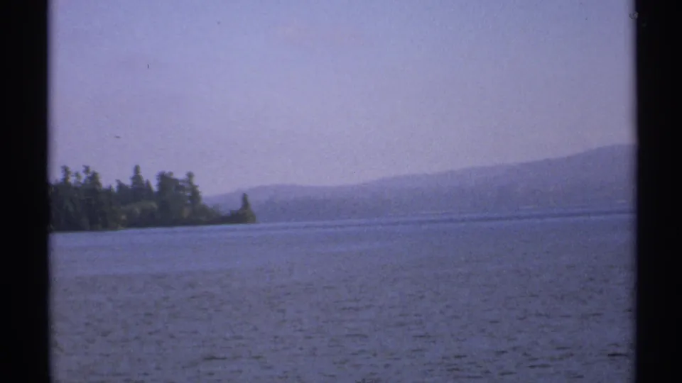 1971 New York 8mm frame showing rippling lake water, a tree-lined shoreline, and distant hazy hills beneath an overcast sky.