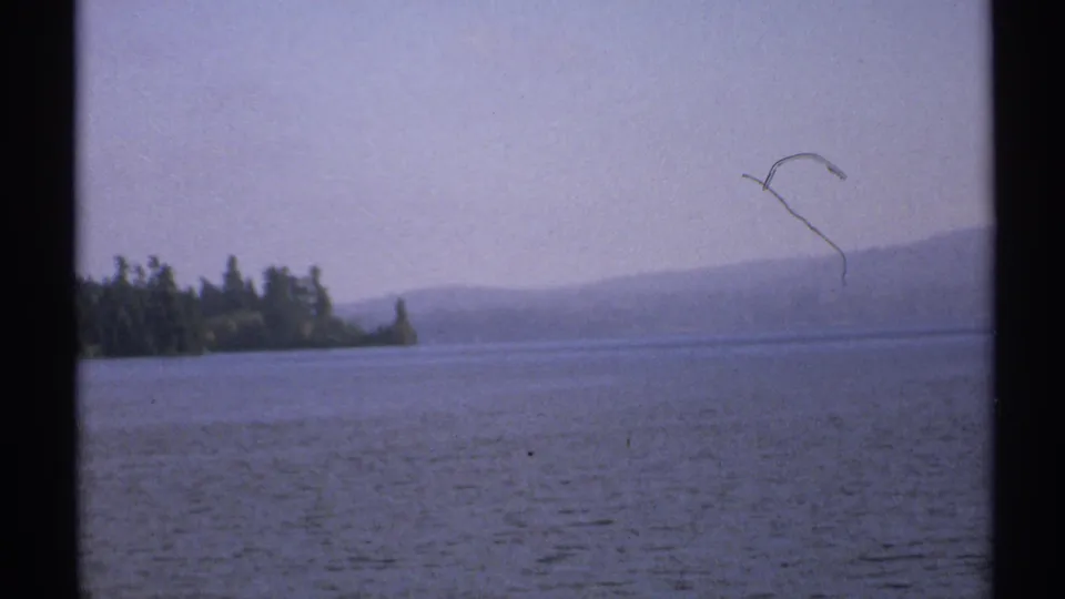 1971 New York 8mm frame of calm lake water and distant wooded shore, with hazy hills and a small dark sky mark or object.