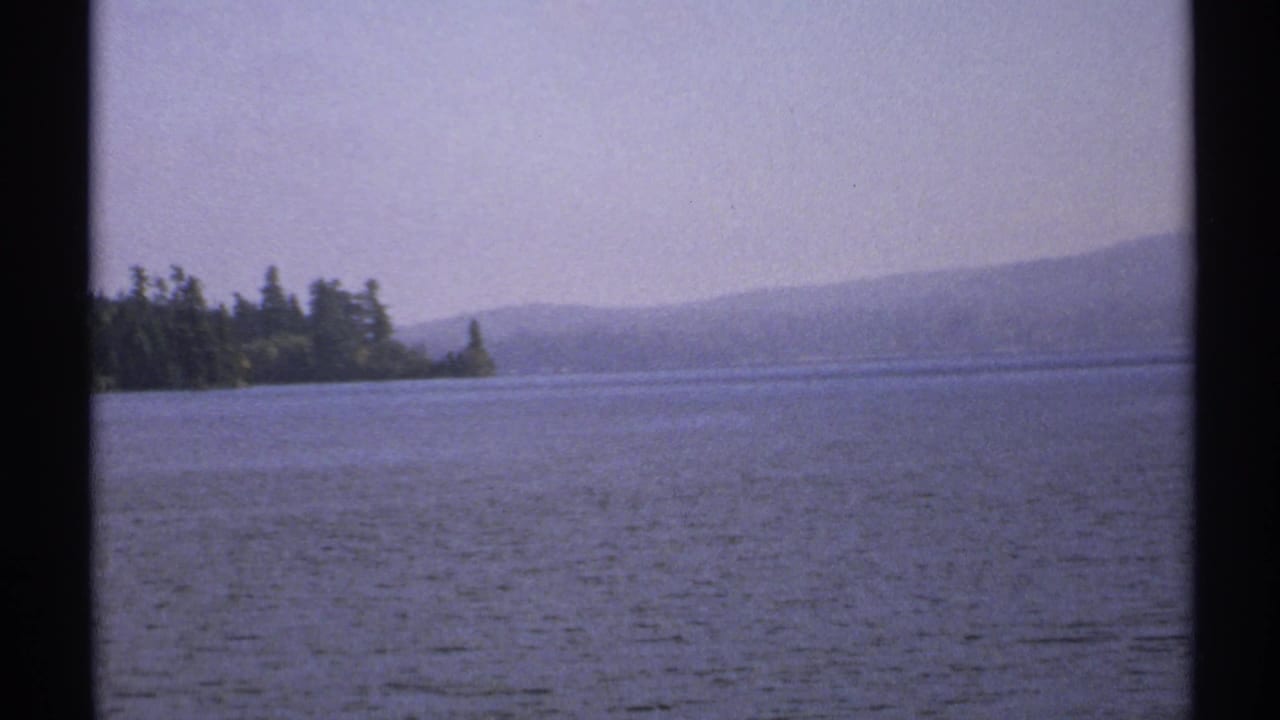 1971 New York 8mm frame presenting a steady lakeside view with rippled water, dark tree line, and low hills softened by haze.