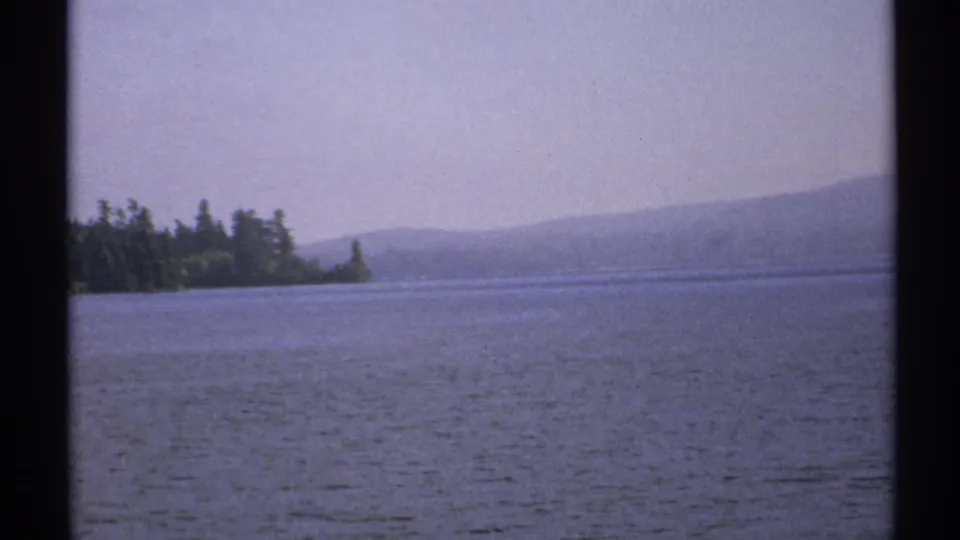1971 New York 8mm frame presenting a steady lakeside view with rippled water, dark tree line, and low hills softened by haze.