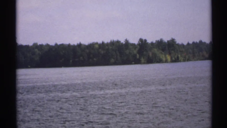 1971 New York 8mm frame showing open lake water and a dark tree-lined shore under a pale sky.