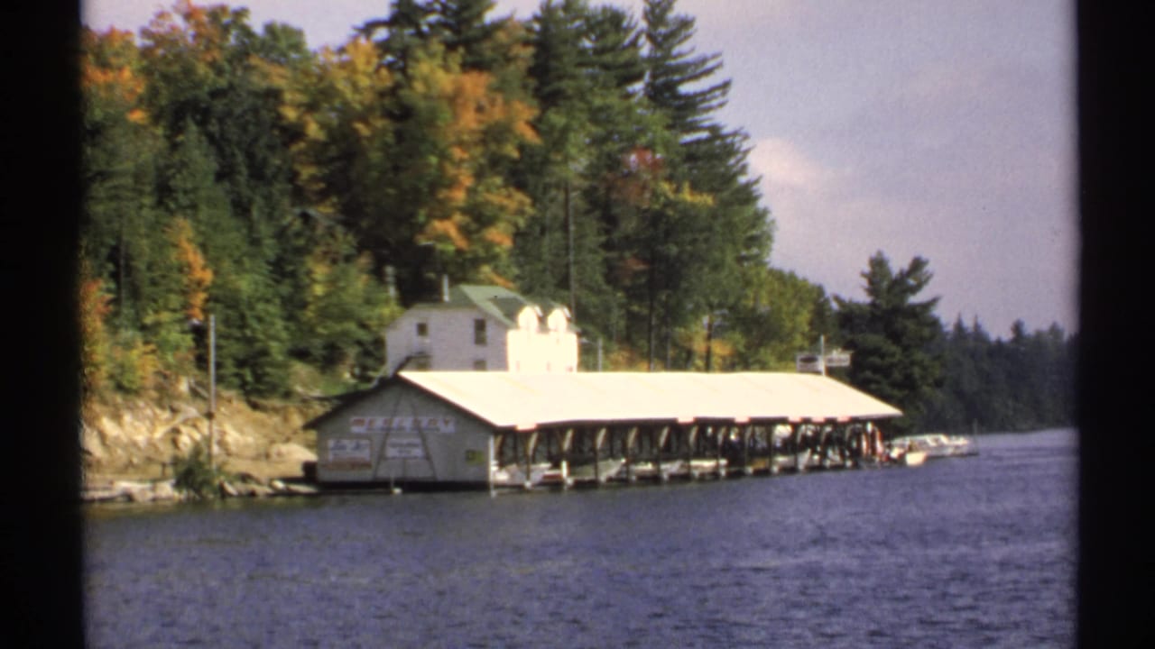 1971 New York 8mm frame showing a white boathouse-like building, long covered dock, boats, lake water, and autumn foliage.