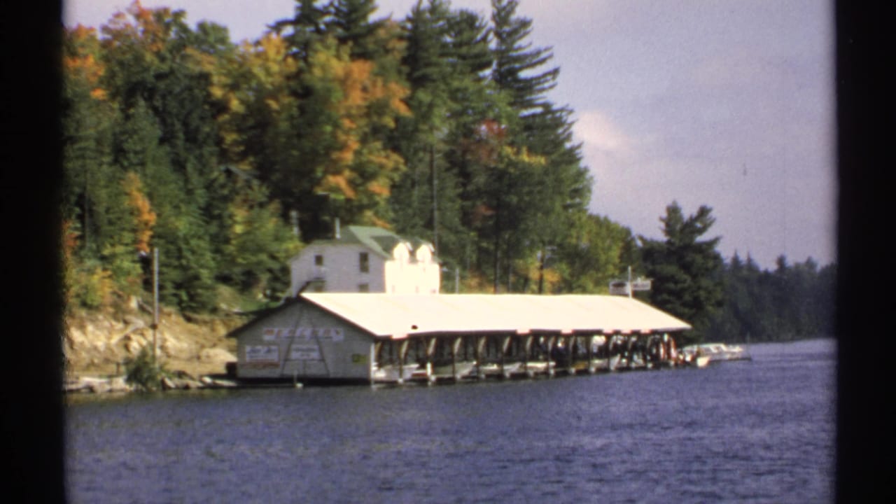 1971 New York 8mm frame showing a lakeside covered dock, small boats, white building, and dense autumn trees from the water.