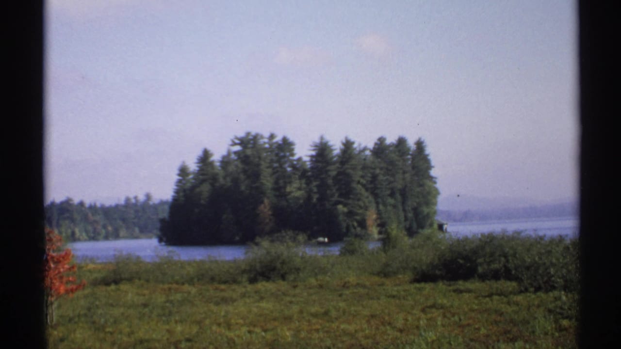 1971 New York 8mm view of a wooded island across a lake, framed by dark window mullions and foreground meadow.