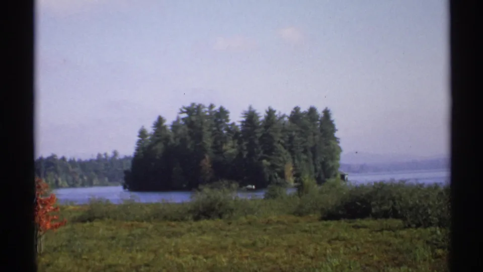 1971 New York 8mm view of a wooded island across a lake, framed by dark window mullions and foreground meadow.