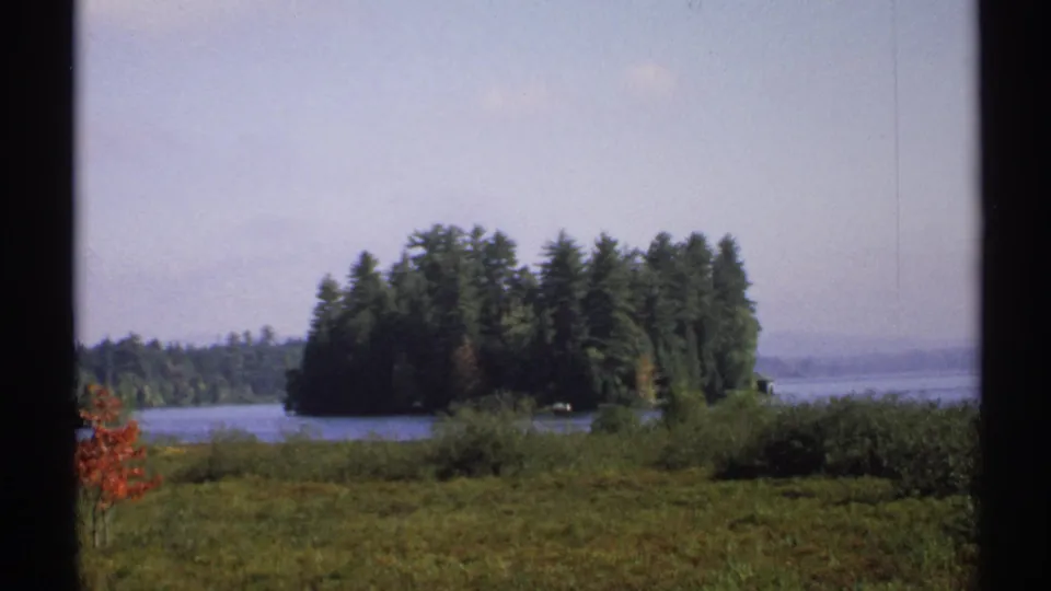 1971 New York home-movie frame showing a lake and dense tree-covered island beyond a grassy foreground.