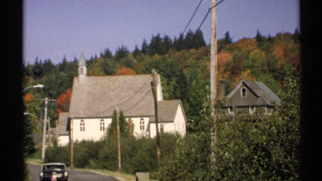 1971 New York street view with a white church, steeple, roadside poles, autumn foliage, shrubs, and a parked car.