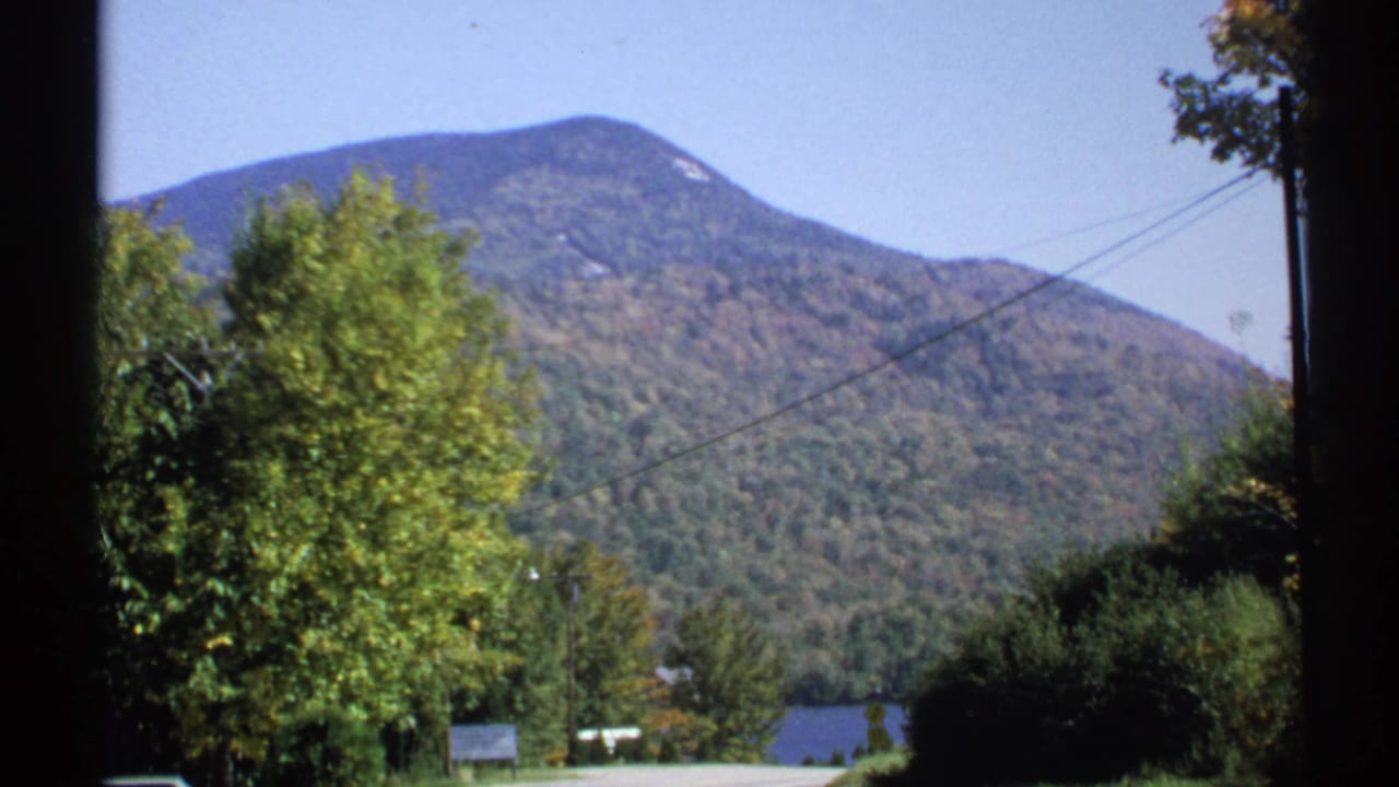 1971 New York 8mm view of a forested mountain beyond roadside trees, utility lines, and a small glimpse of water.