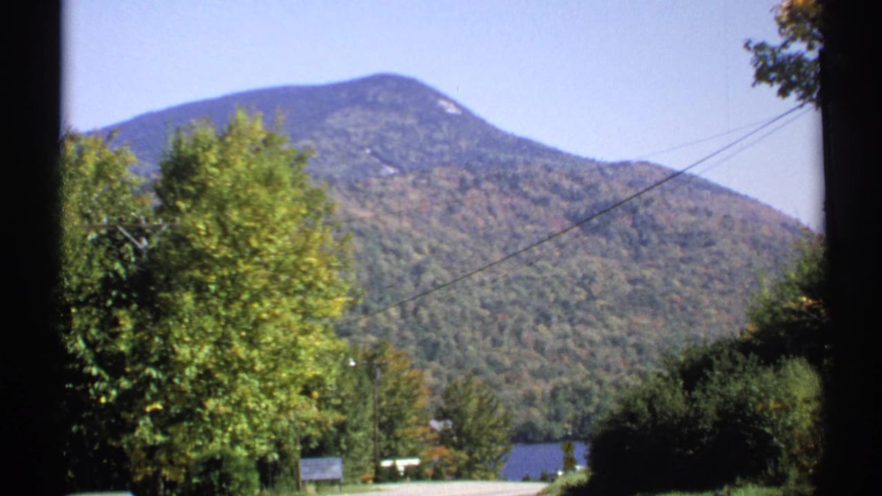 1971 New York 8mm frame of a forested mountain, roadside trees, power lines, and a distant lake.