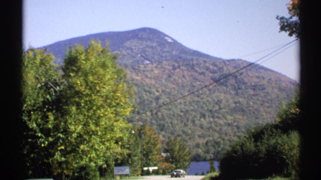 1971 New York 8mm view of a forested peak, lakeshore trees, power lines, and a small car in the distance.
