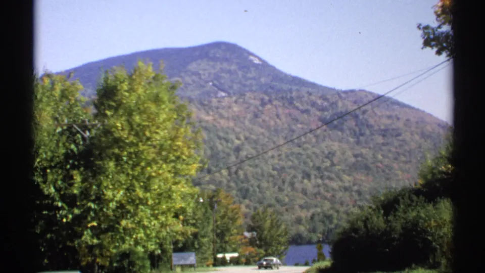 1971 New York 8mm view of a forested peak, lakeshore trees, power lines, and a small car in the distance.