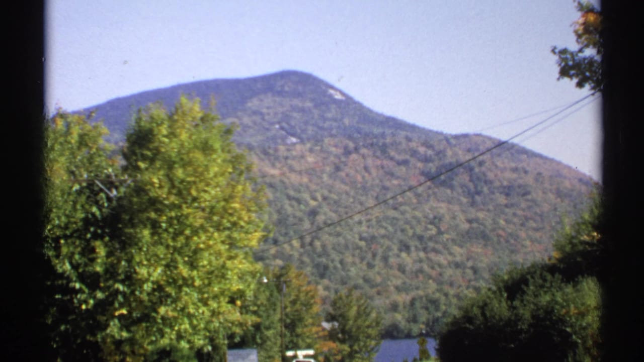 1971 New York 8mm frame showing a forested mountain, roadside foliage, power lines, and a partial lake view.