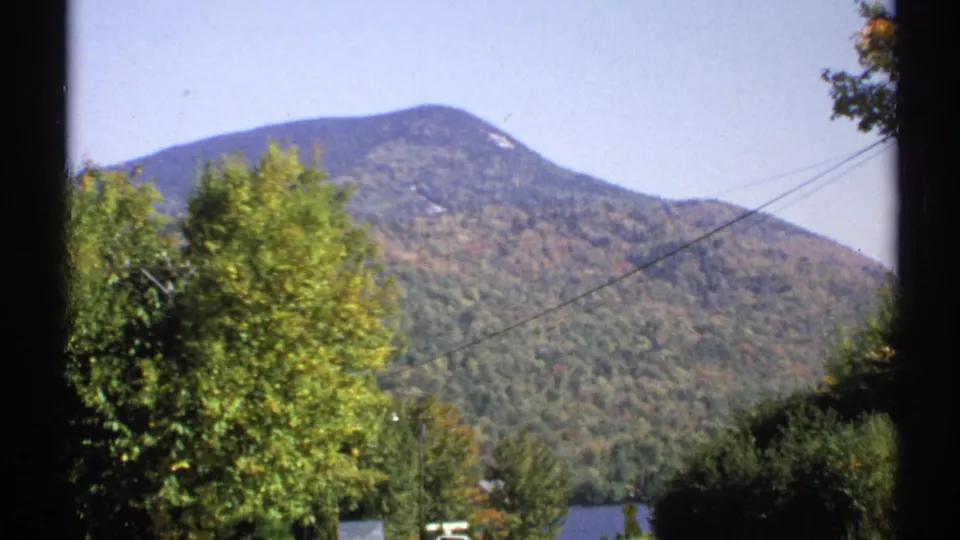 1971 New York 8mm frame showing a forested mountain, roadside foliage, power lines, and a partial lake view.