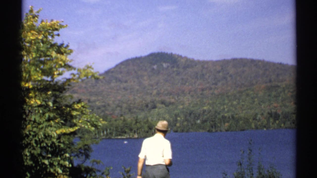 1971 New York 8mm lakeside frame with a man in a light shirt and hat looking toward a forested mountain.
