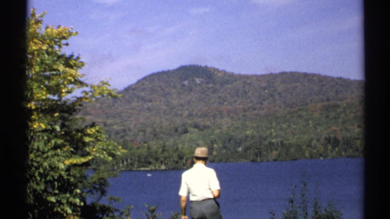 1971 New York 8mm frame of a man seen from behind at a lakeside viewpoint, looking toward wooded hills under a blue sky.