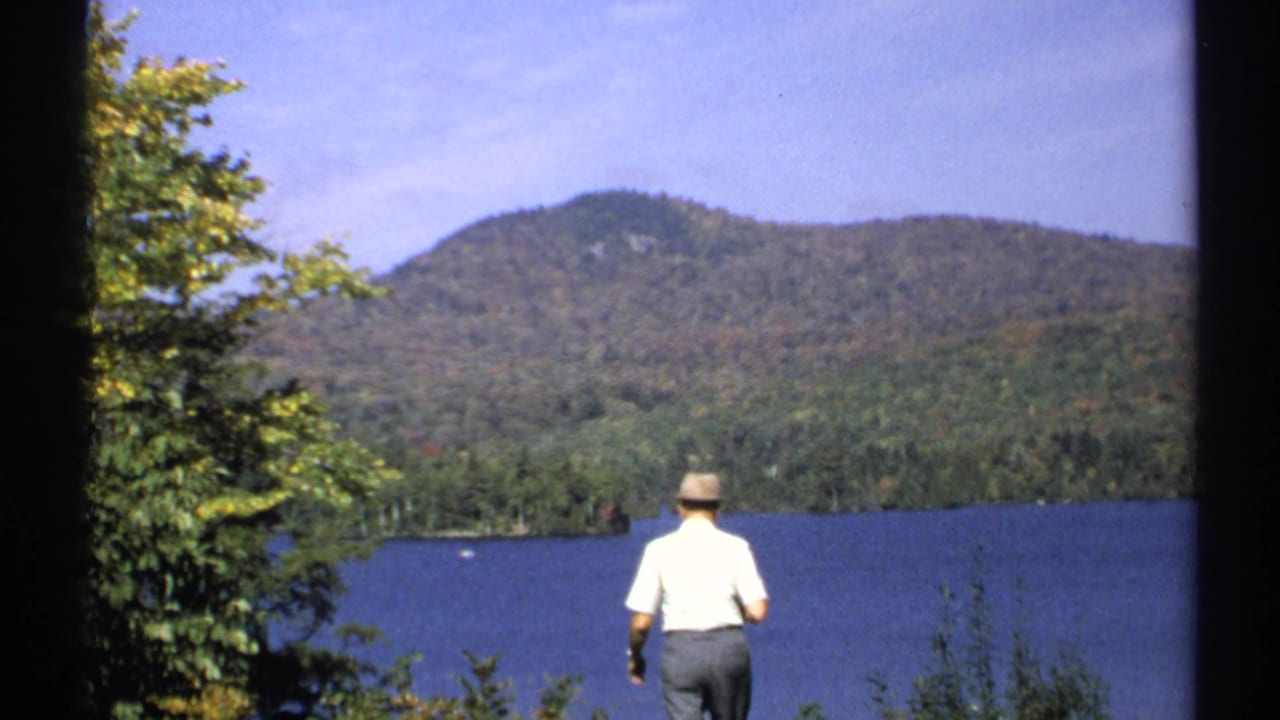 Man walks or stands near the lake with wooded hills beyond.