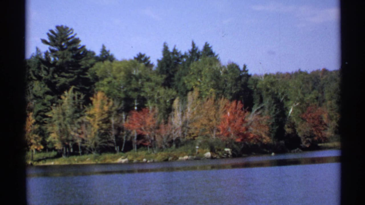 1971 New York 8mm frame looking across lake water toward a forested shoreline with fall foliage.